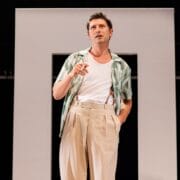 A man in a patterned shirt and beige trousers stands in front of a rectangular frame, holding a cigarette and looking upward, evoking the moody atmosphere of a psychological thriller like The Talented Mr. Ripley.