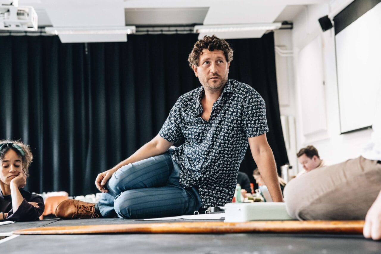 A man in a patterned shirt, evoking the intrigue of a psychological thriller like The Talented Mr. Ripley, sits cross-legged on a table in a room with black curtains, surrounded by people and papers.