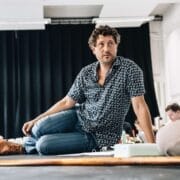 A man in a patterned shirt, evoking the intrigue of a psychological thriller like The Talented Mr. Ripley, sits cross-legged on a table in a room with black curtains, surrounded by people and papers.