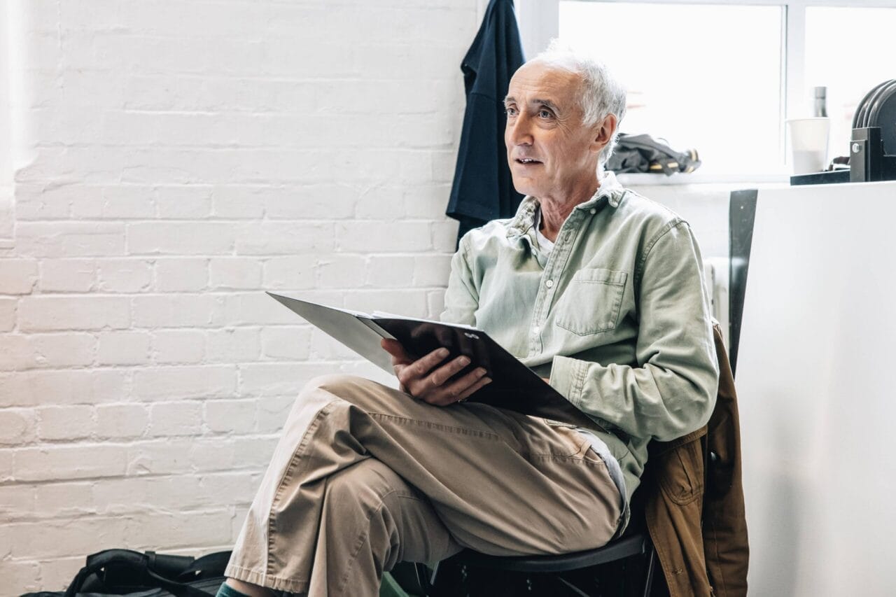 Older man sitting indoors holding a folder, wearing a light green shirt and beige pants, with a jacket draped over the chair—a scene reminiscent of The Talented Mr. Ripley, with casual items in the background.