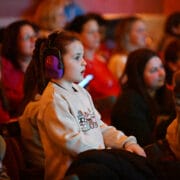 A young girl with headphones and a beige sweater attentively watches an event in a dimly lit auditorium, surrounded by other seated attendees.
