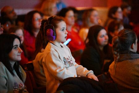 A young girl with headphones and a beige sweater attentively watches an event in a dimly lit auditorium, surrounded by other seated attendees.