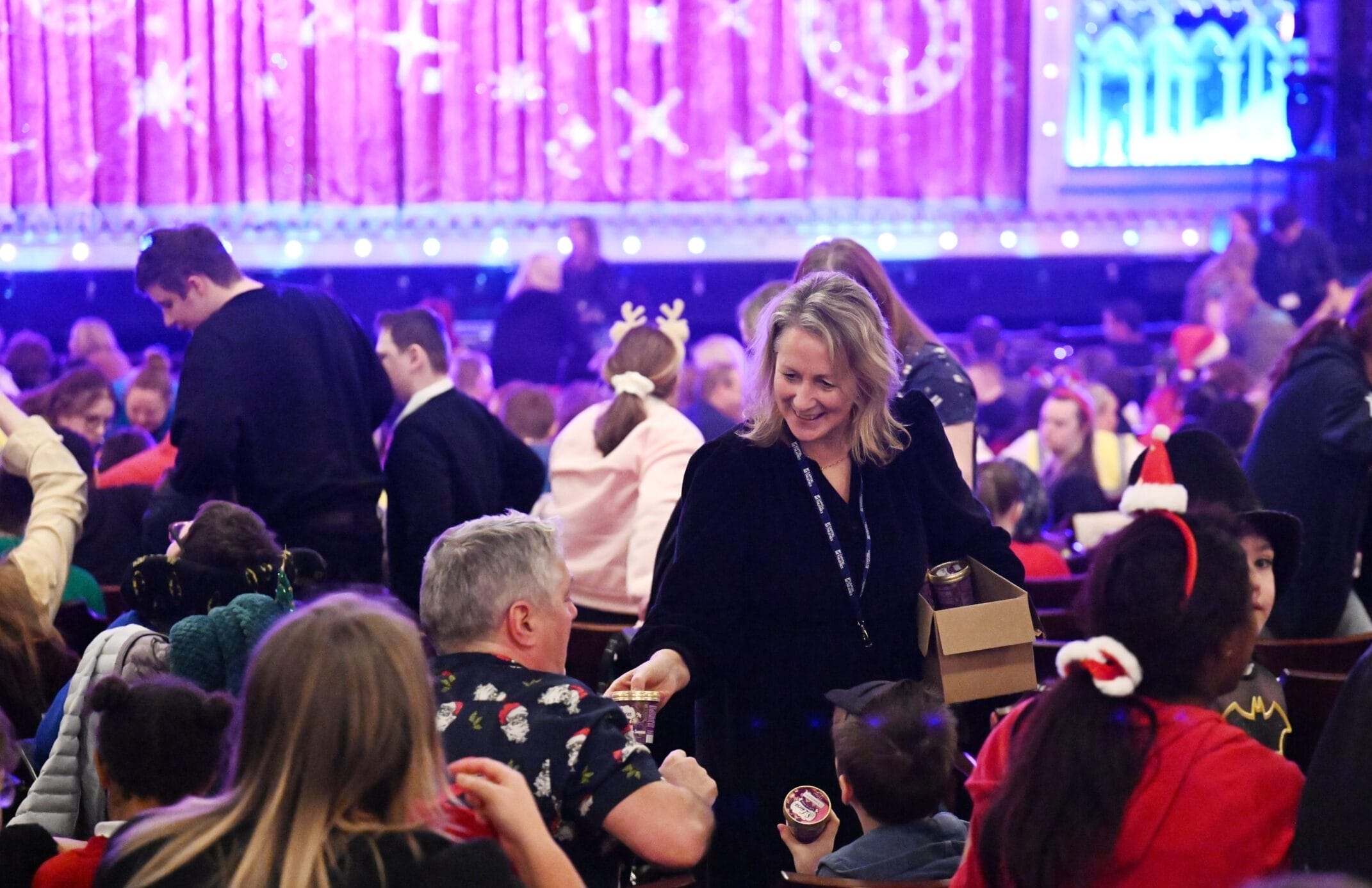 A woman hands out snacks to seated audience members in a crowded theater decorated with holiday lights and festive decorations.