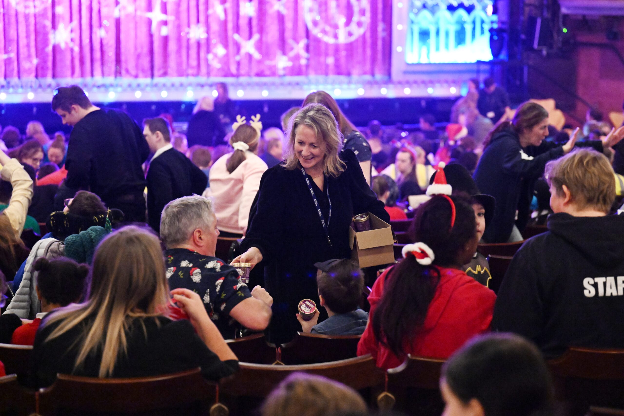 A woman hands out snacks to seated audience members in a crowded theater decorated with holiday lights and festive decorations.