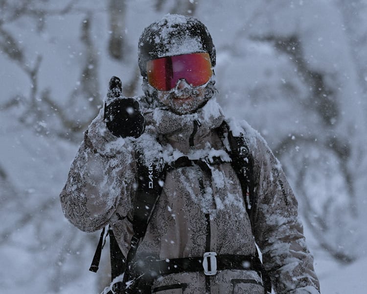 A person in snow gear and goggles stands outdoors in heavy snowfall, giving a thumbs up while supporting the Blue Programme.
