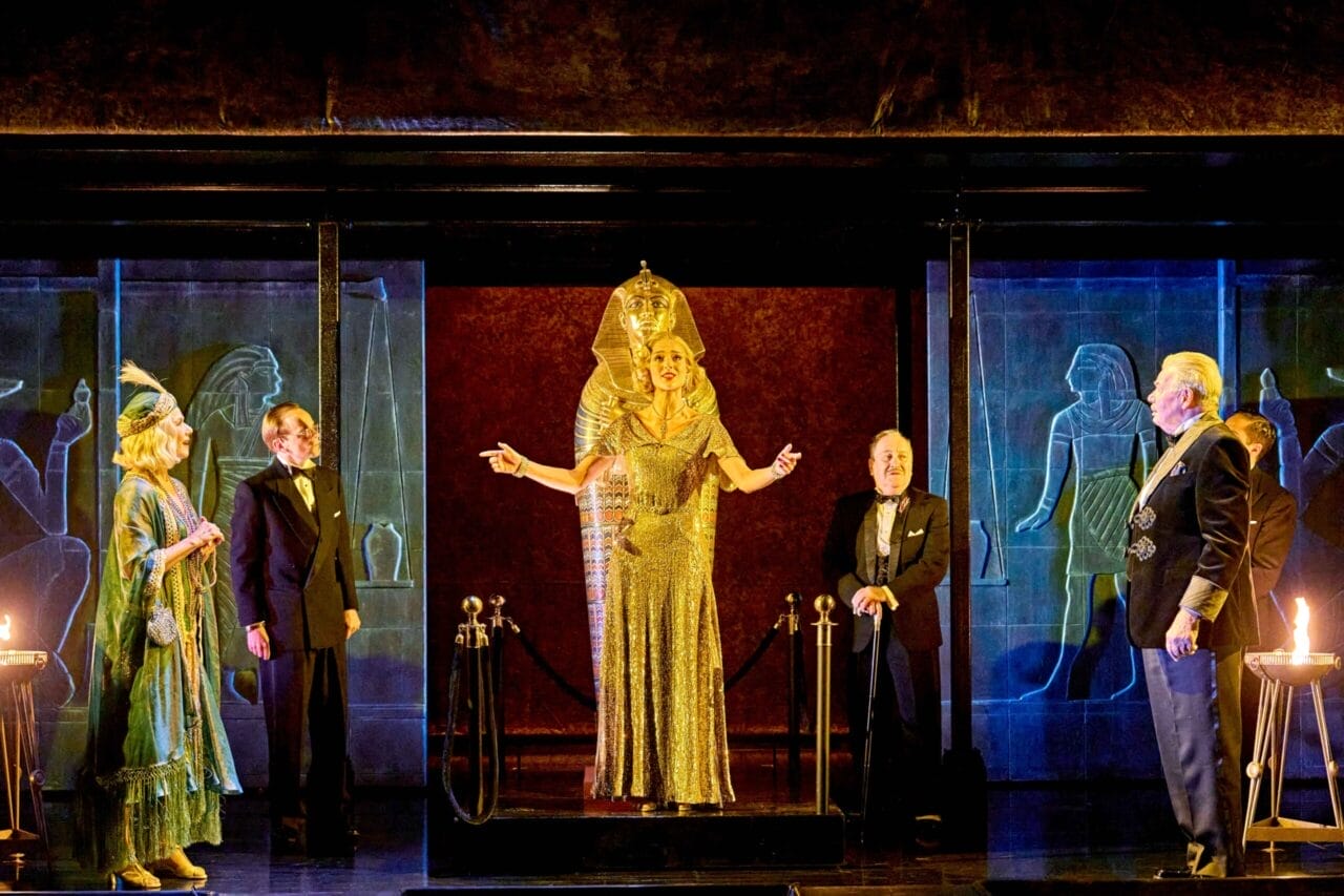 Five people stand on stage in formal and period costumes, with one person dressed as an Egyptian pharaoh in gold at the center, surrounded by an ancient Egyptian-themed set inspired by Death on the Nile.