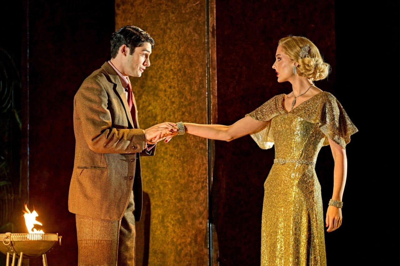 A man in a brown suit holds the hand of a woman in a gold dress. Reminiscent of "Death on the Nile," they face each other on a dimly lit stage, with a small fire flickering mysteriously in the background.