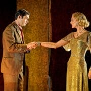 A man in a brown suit holds the hand of a woman in a gold dress. Reminiscent of "Death on the Nile," they face each other on a dimly lit stage, with a small fire flickering mysteriously in the background.