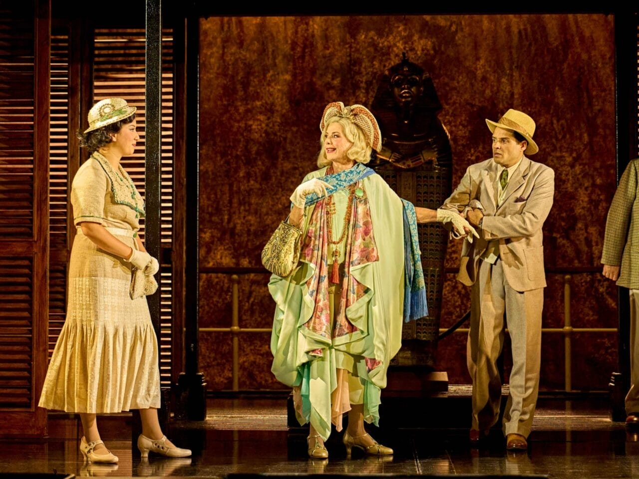 Three actors in period costumes stand on stage, one in a green and floral outfit. A sarcophagus looms in the background, evoking the intrigue of Death on the Nile with its Egyptian or archaeological theme.