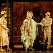 Three actors in period costumes stand on stage, one in a green and floral outfit. A sarcophagus looms in the background, evoking the intrigue of Death on the Nile with its Egyptian or archaeological theme.