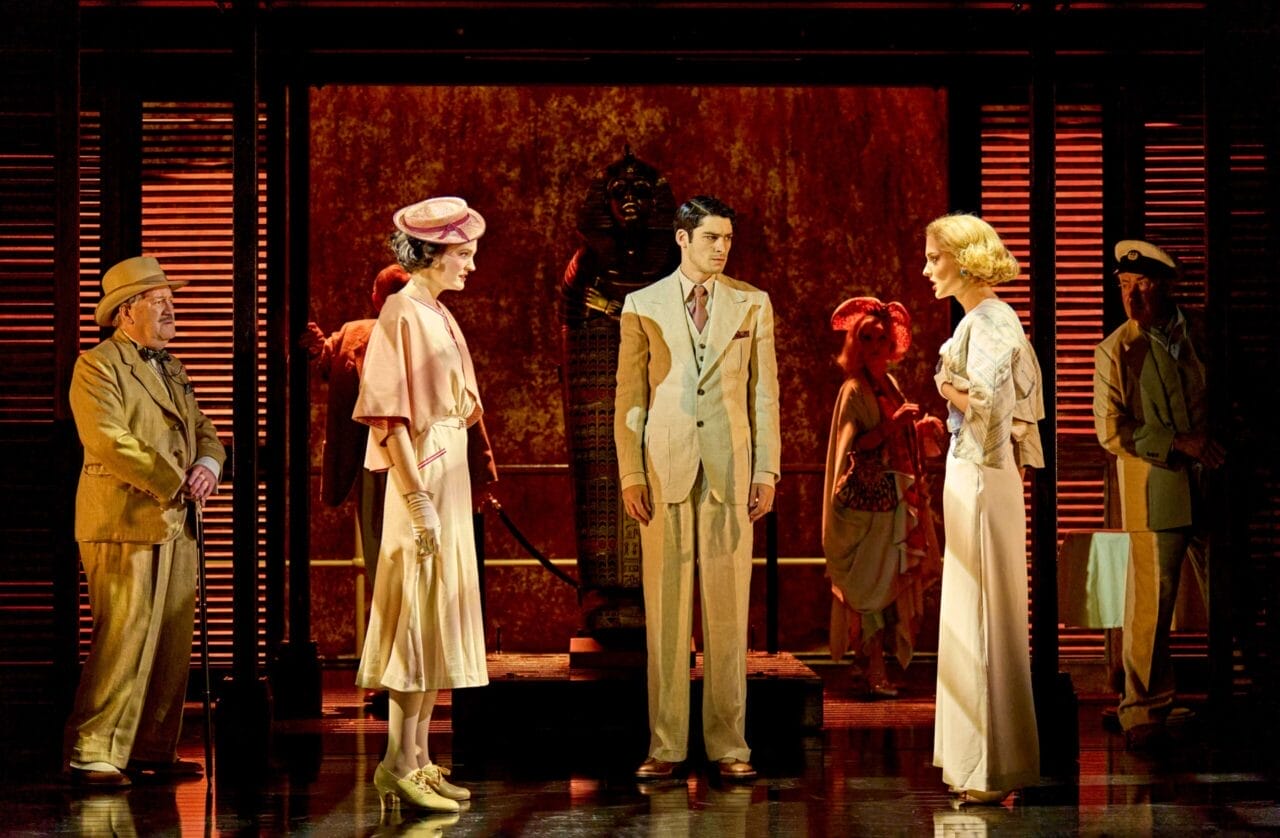 Actors in period costumes stand on a stage set resembling ancient Egypt, with a statue in the background and dramatic red lighting evoking the suspenseful ambiance of Death on the Nile.