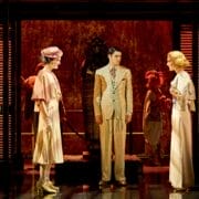 Actors in period costumes stand on a stage set resembling ancient Egypt, with a statue in the background and dramatic red lighting evoking the suspenseful ambiance of Death on the Nile.