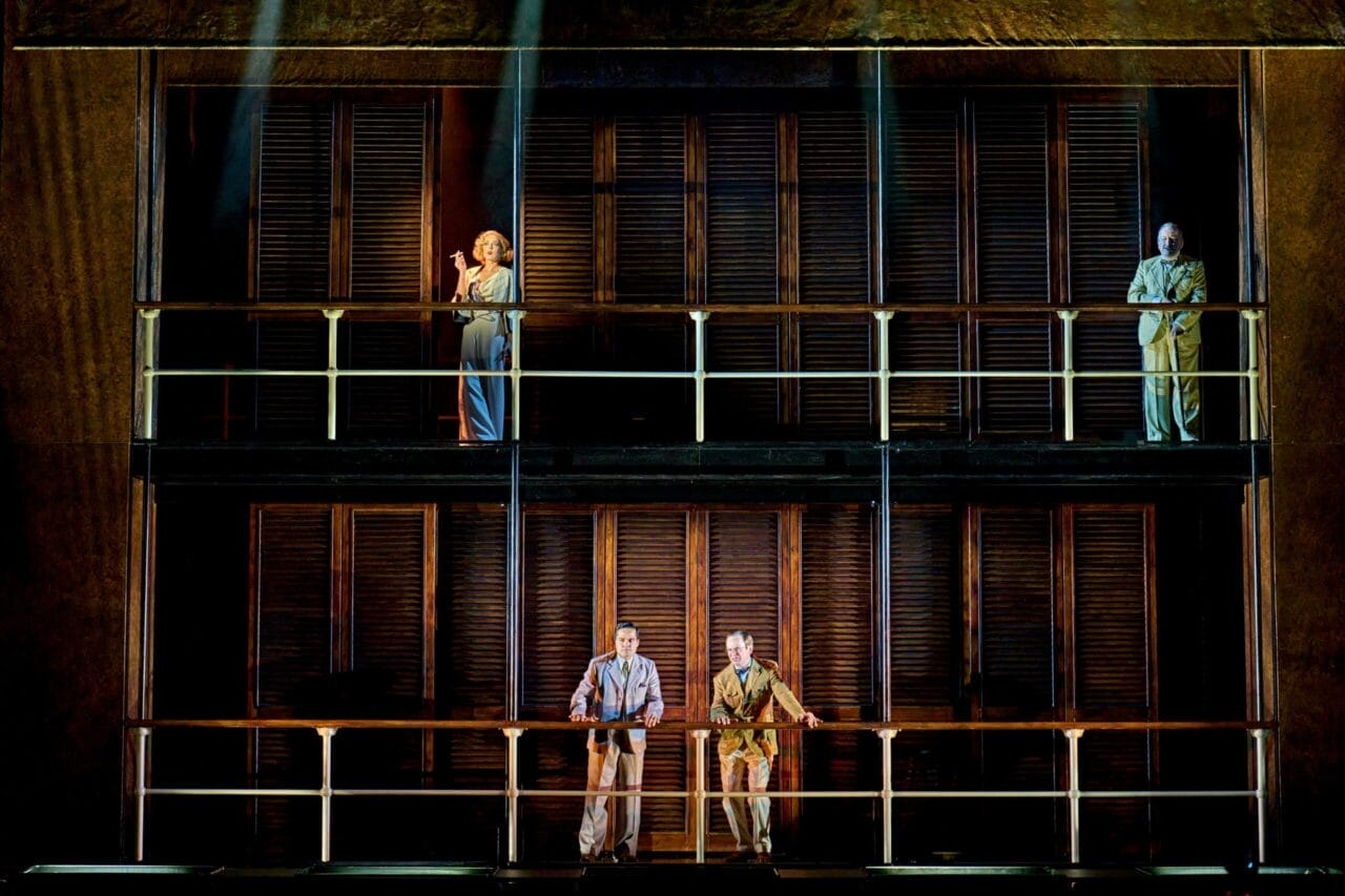 Four people stand on two levels of a stage set with wooden panels and railings, dramatically lit as though poised for a scene from Death on the Nile.