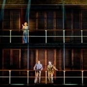 Four people stand on two levels of a stage set with wooden panels and railings, dramatically lit as though poised for a scene from Death on the Nile.