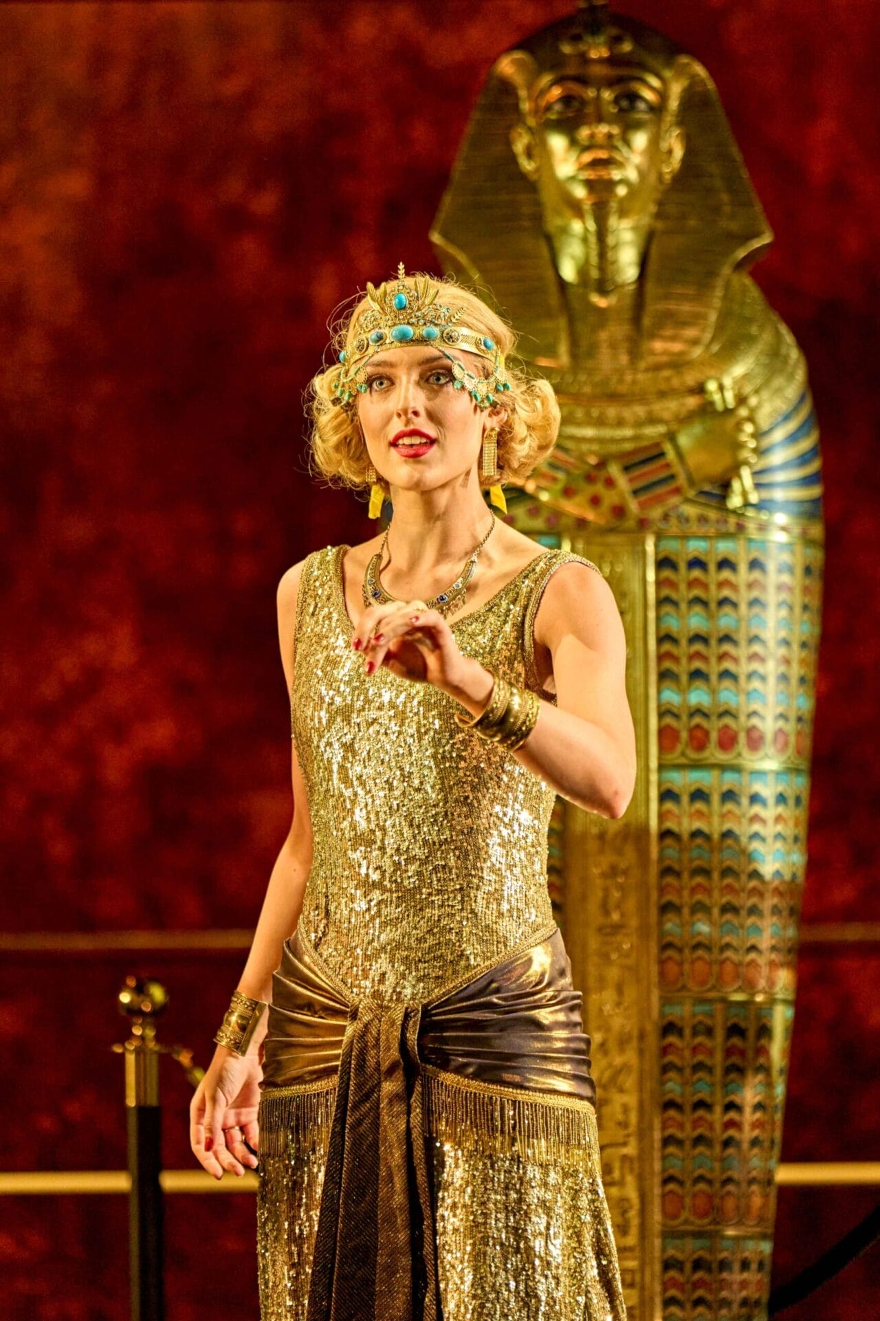 A woman in a gold, 1920s-style dress and jeweled headpiece stands before an ornate Egyptian sarcophagus, evoking the intrigue of Death on the Nile, set against a rich red backdrop.
