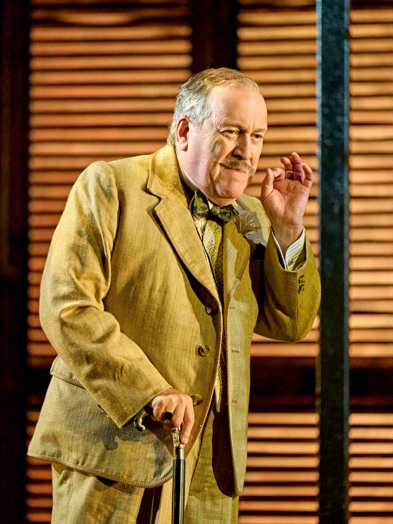 An older man in a tan suit holds a cane and gestures with his right hand in front of wooden shutters, evoking the elegance of Death on the Nile.