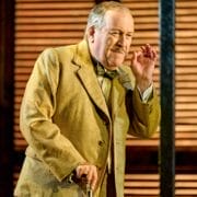 An older man in a tan suit holds a cane and gestures with his right hand in front of wooden shutters, evoking the elegance of Death on the Nile.