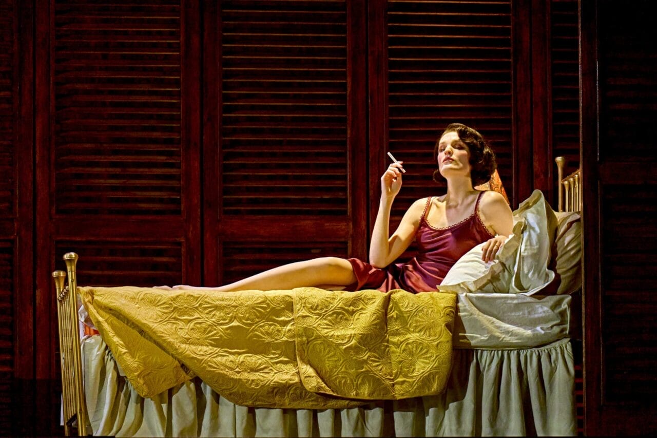A woman in a maroon slip dress reclines on a bed with yellow bedding, holding a cigarette, evoking Death on the Nile glamour against a backdrop of dark wooden shutters.