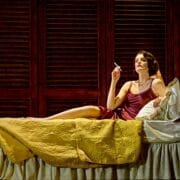 A woman in a maroon slip dress reclines on a bed with yellow bedding, holding a cigarette, evoking Death on the Nile glamour against a backdrop of dark wooden shutters.