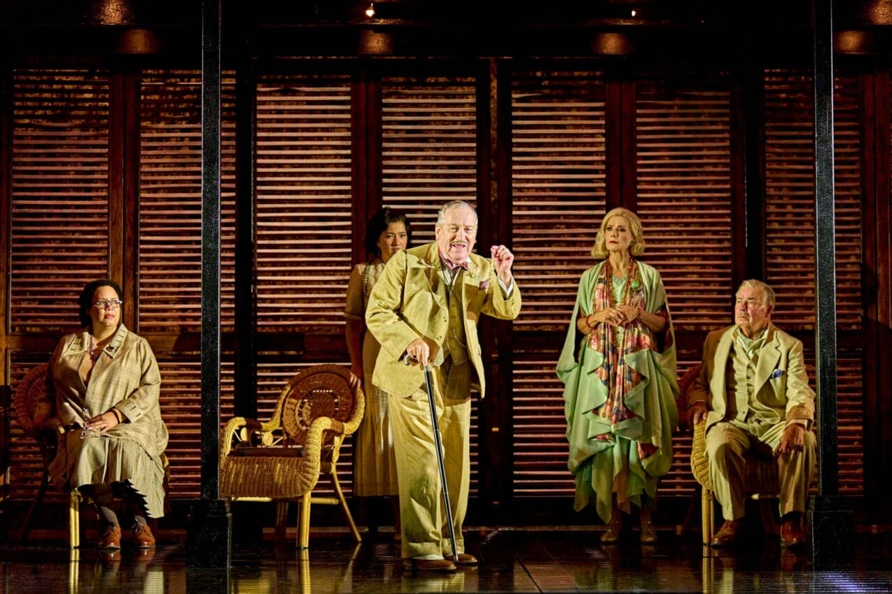 Five actors in period costumes perform on a stage with wooden shutters and wicker chairs, evoking a dramatic scene inspired by Death on the Nile in a vintage setting.