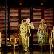 Five actors in period costumes perform on a stage with wooden shutters and wicker chairs, evoking a dramatic scene inspired by Death on the Nile in a vintage setting.