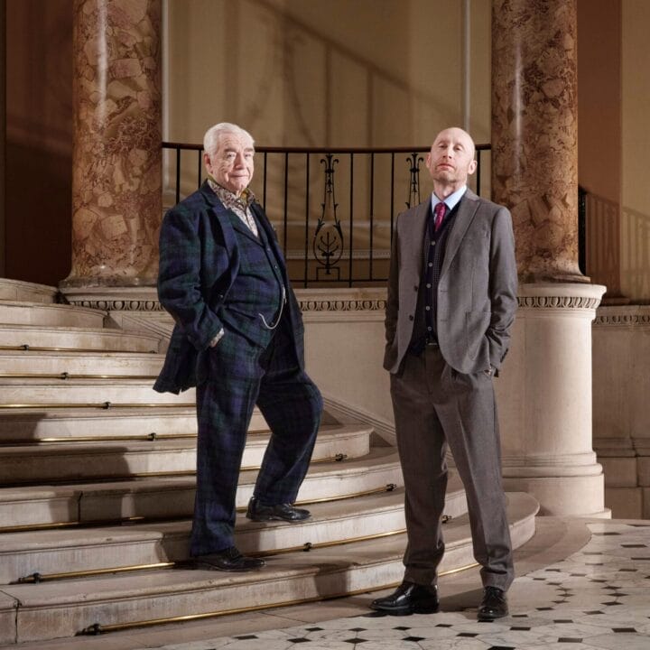 Two men in suits, Brian Cox on the left and Sandy Grierson on the right, stand on a marble staircase in an ornate interior with tall columns and patterned floor tiles.
