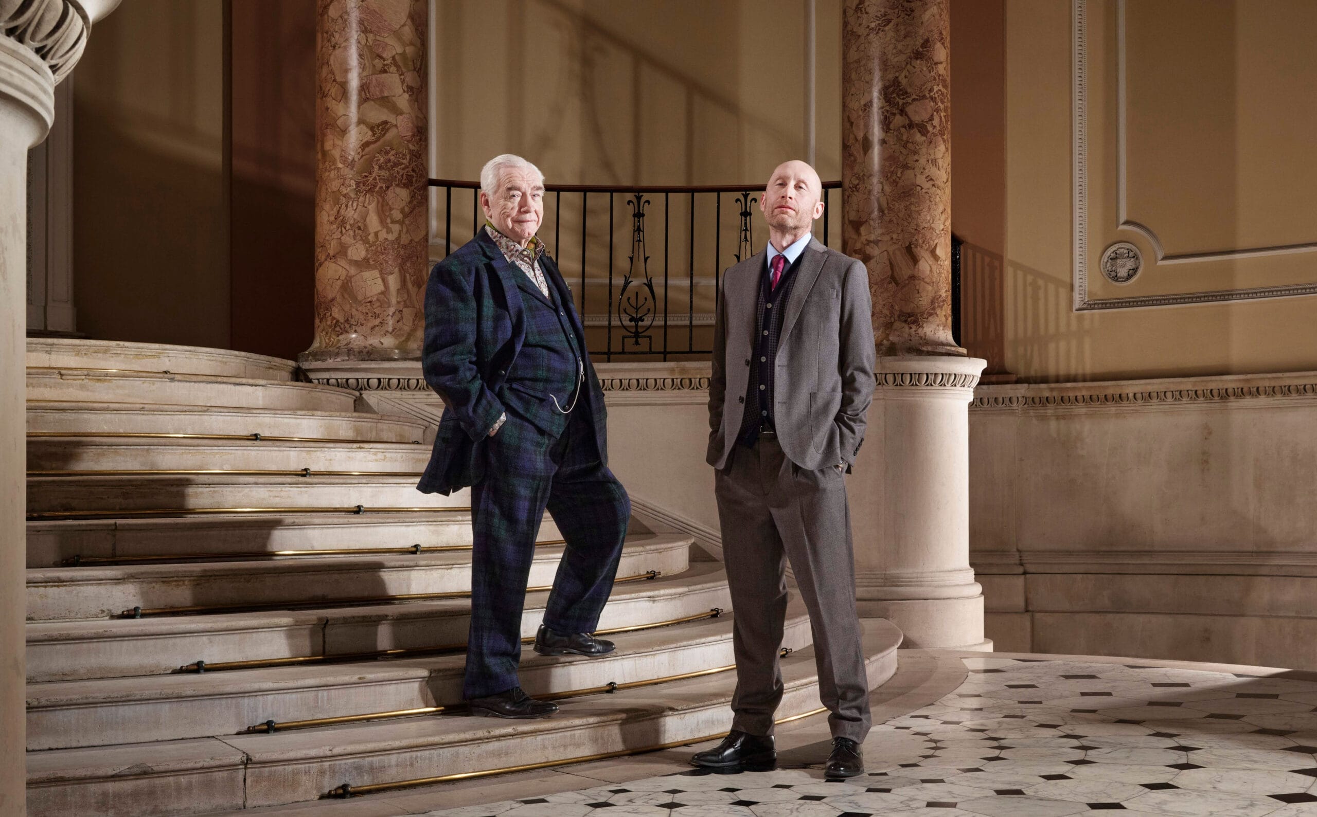 Two men in suits, Brian Cox on the left and Sandy Grierson on the right, stand on a marble staircase in an ornate interior with tall columns and patterned floor tiles.
