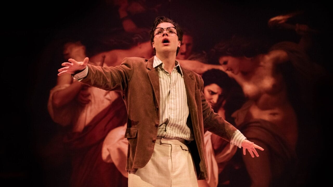 A person wearing glasses, a brown jacket, and light pants stands on stage with arms outstretched, evoking The Talented Mr. Ripley in front of a dramatic painting backdrop.