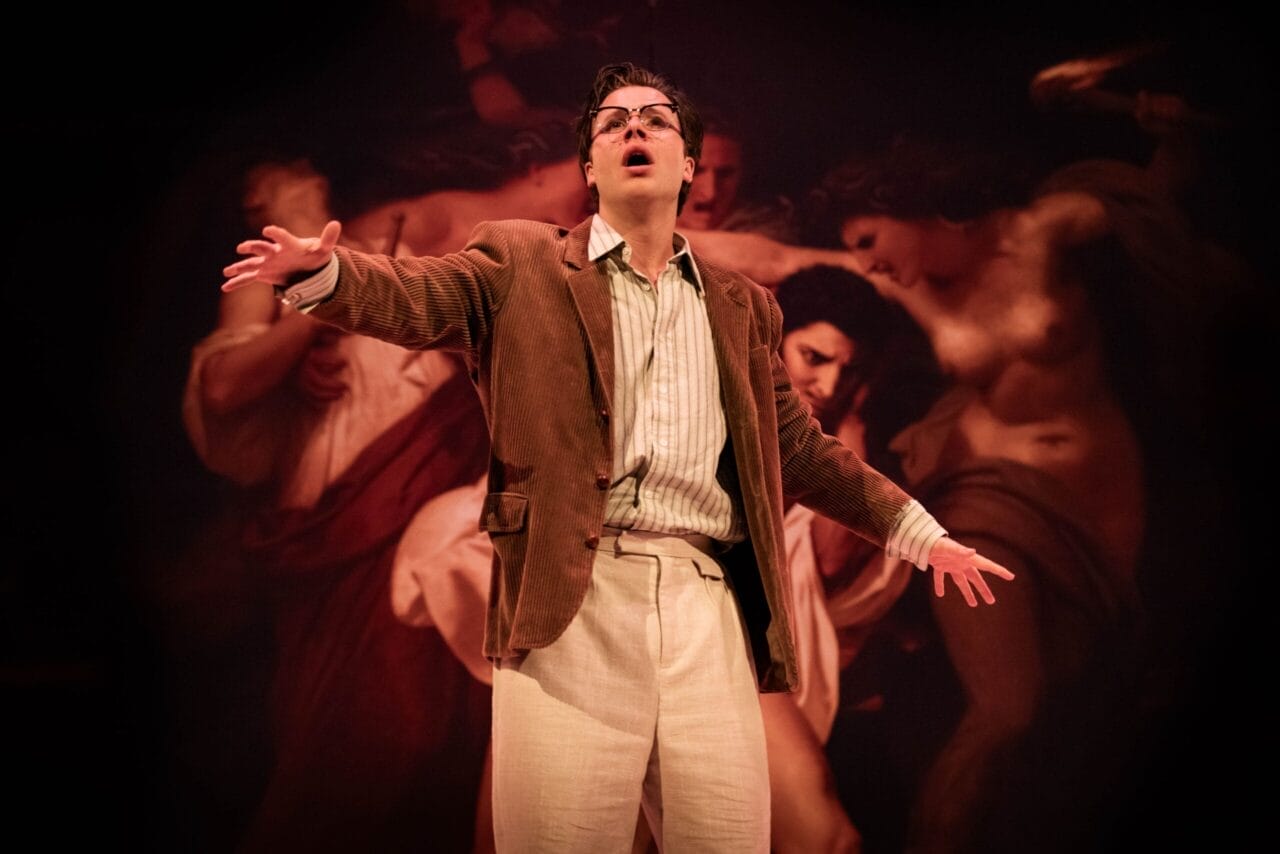 A person wearing glasses, a brown jacket, and light pants stands on stage with arms outstretched, evoking The Talented Mr. Ripley in front of a dramatic painting backdrop.