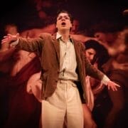 A person wearing glasses, a brown jacket, and light pants stands on stage with arms outstretched, evoking The Talented Mr. Ripley in front of a dramatic painting backdrop.