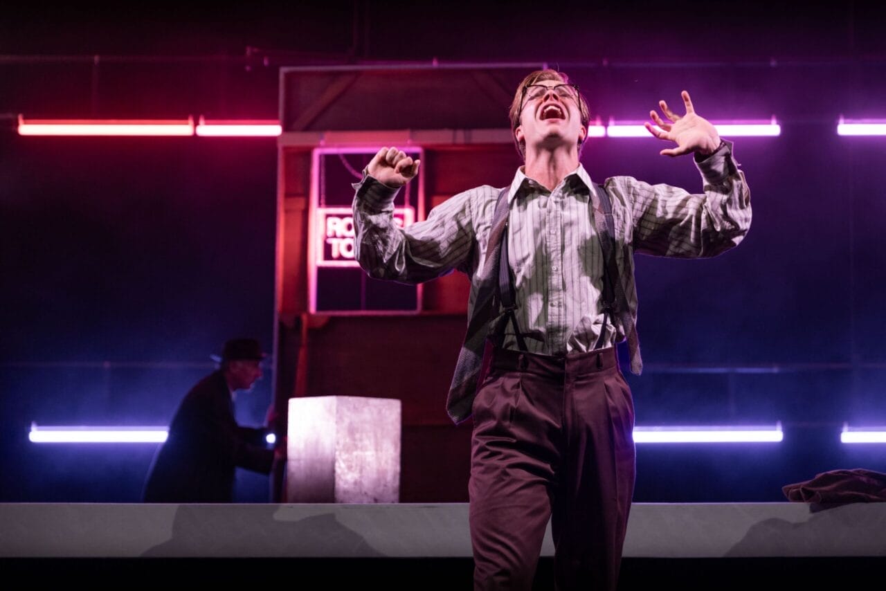 A person in glasses and suspenders performs dramatically on stage under neon lights, evoking The Talented Mr. Ripley, while another figure lingers in the background by a counter.