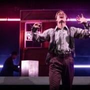 A person in glasses and suspenders performs dramatically on stage under neon lights, evoking The Talented Mr. Ripley, while another figure lingers in the background by a counter.