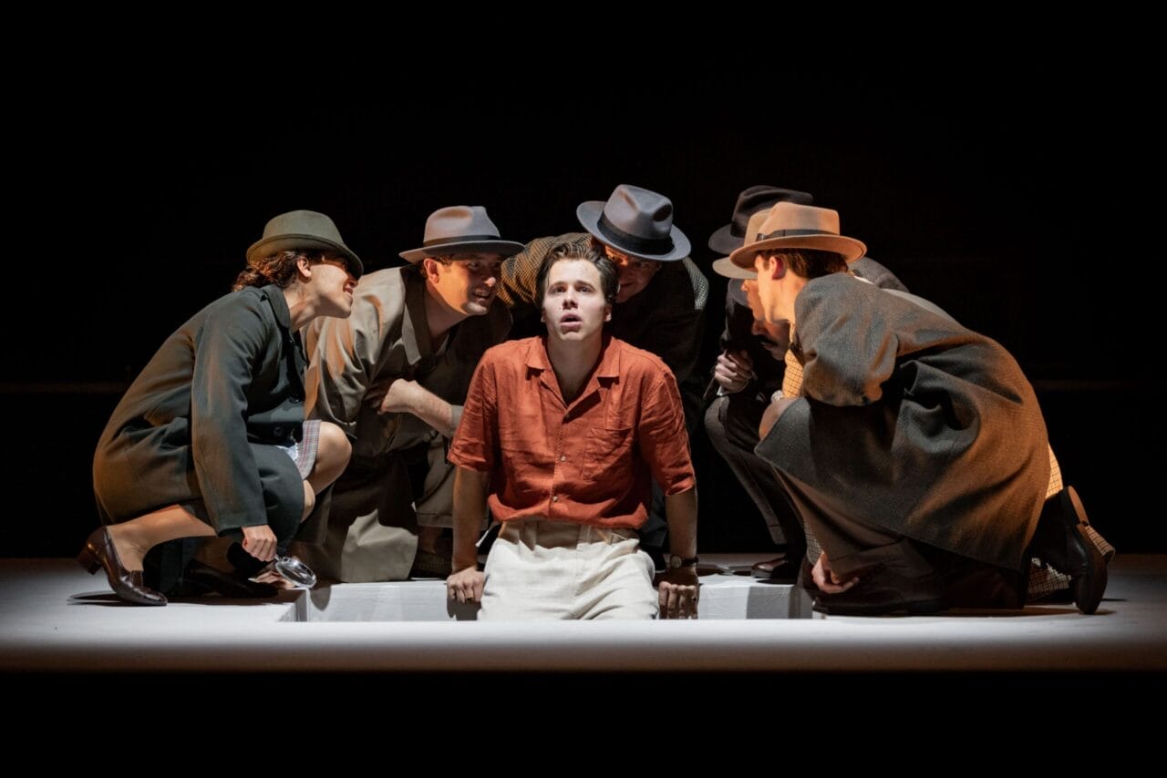 A man in an orange shirt and beige pants sits surrounded by four people in hats and trench coats, leaning in toward him on a dimly lit stage, evoking the suspenseful atmosphere of The Talented Mr. Ripley.