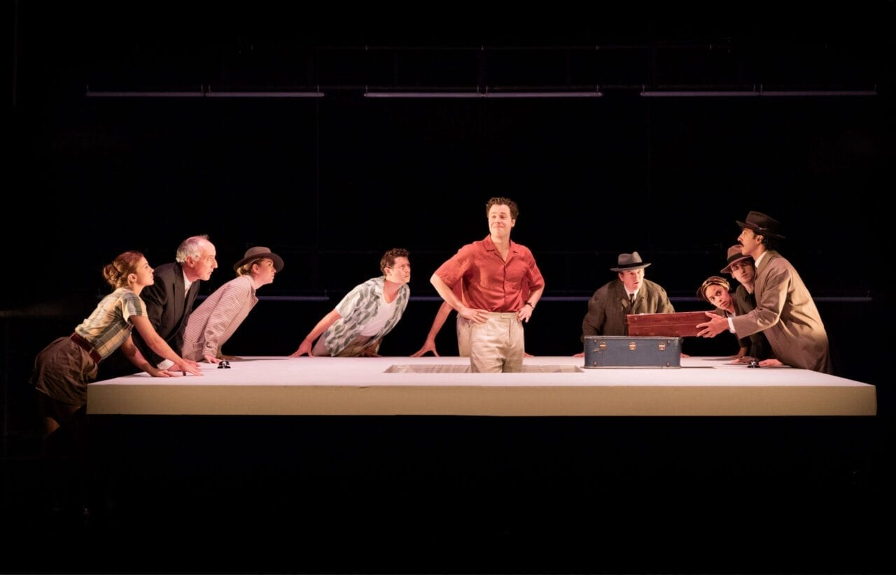 Eight people stand or lean around a large white table with a blue trunk on it, while one person stands in the center, facing forward—a scene reminiscent of The Talented Mr. Ripley. The background is dark.