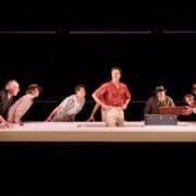 Eight people stand or lean around a large white table with a blue trunk on it, while one person stands in the center, facing forward—a scene reminiscent of The Talented Mr. Ripley. The background is dark.