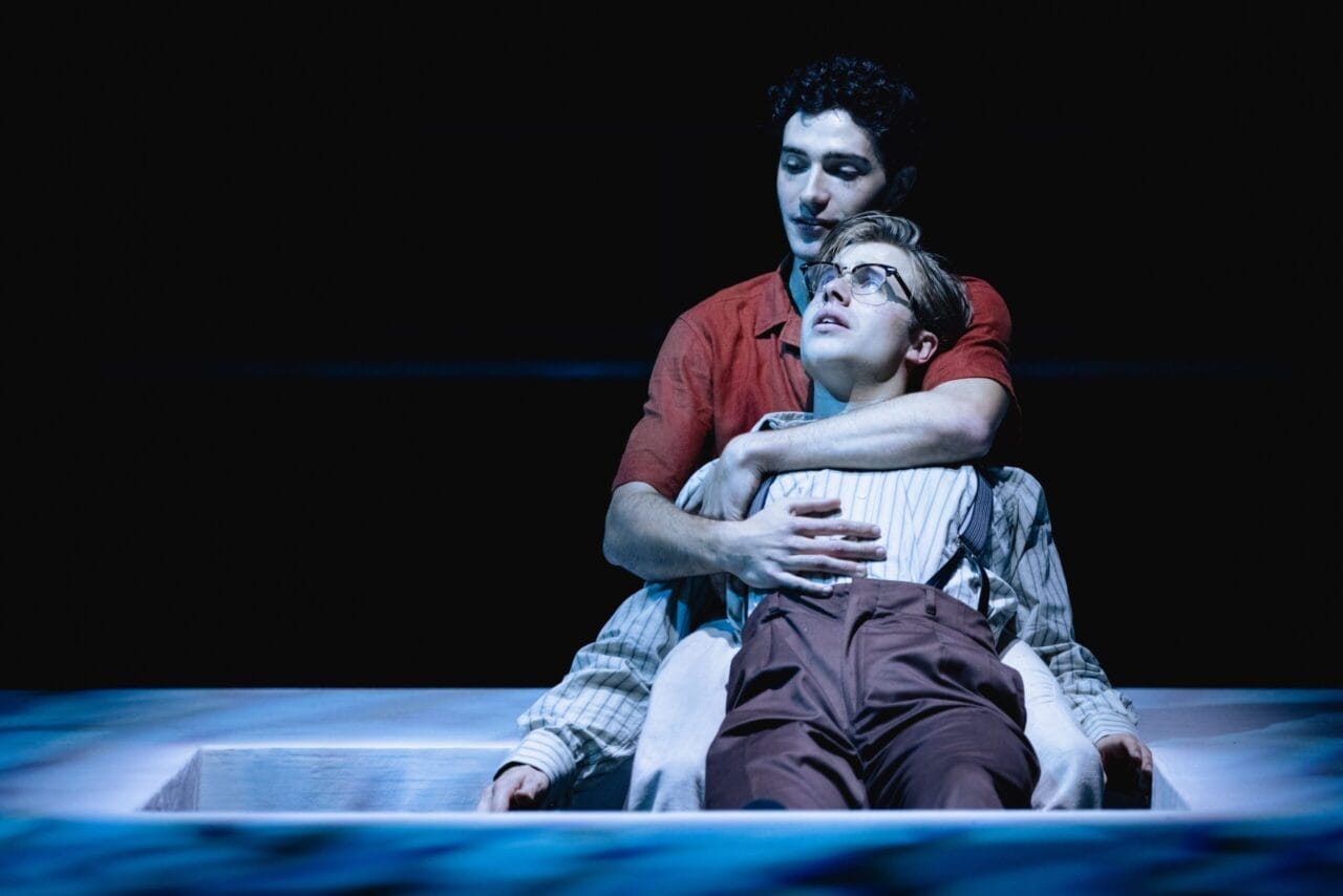 Two people sit closely together on stage, with one person leaning back against the other, who has their arms wrapped around them, evoking an intimate moment reminiscent of The Talented Mr. Ripley, all under dramatic lighting.