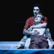 Two people sit closely together on stage, with one person leaning back against the other, who has their arms wrapped around them, evoking an intimate moment reminiscent of The Talented Mr. Ripley, all under dramatic lighting.
