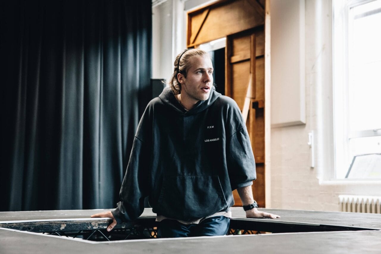 A person with long hair in a dark hoodie leans on a table, looking to the side, in a room with black curtains and natural light from a window—evoking the intrigue of The Talented Mr. Ripley.