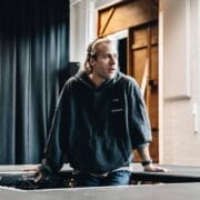 A person with long hair in a dark hoodie leans on a table, looking to the side, in a room with black curtains and natural light from a window—evoking the intrigue of The Talented Mr. Ripley.