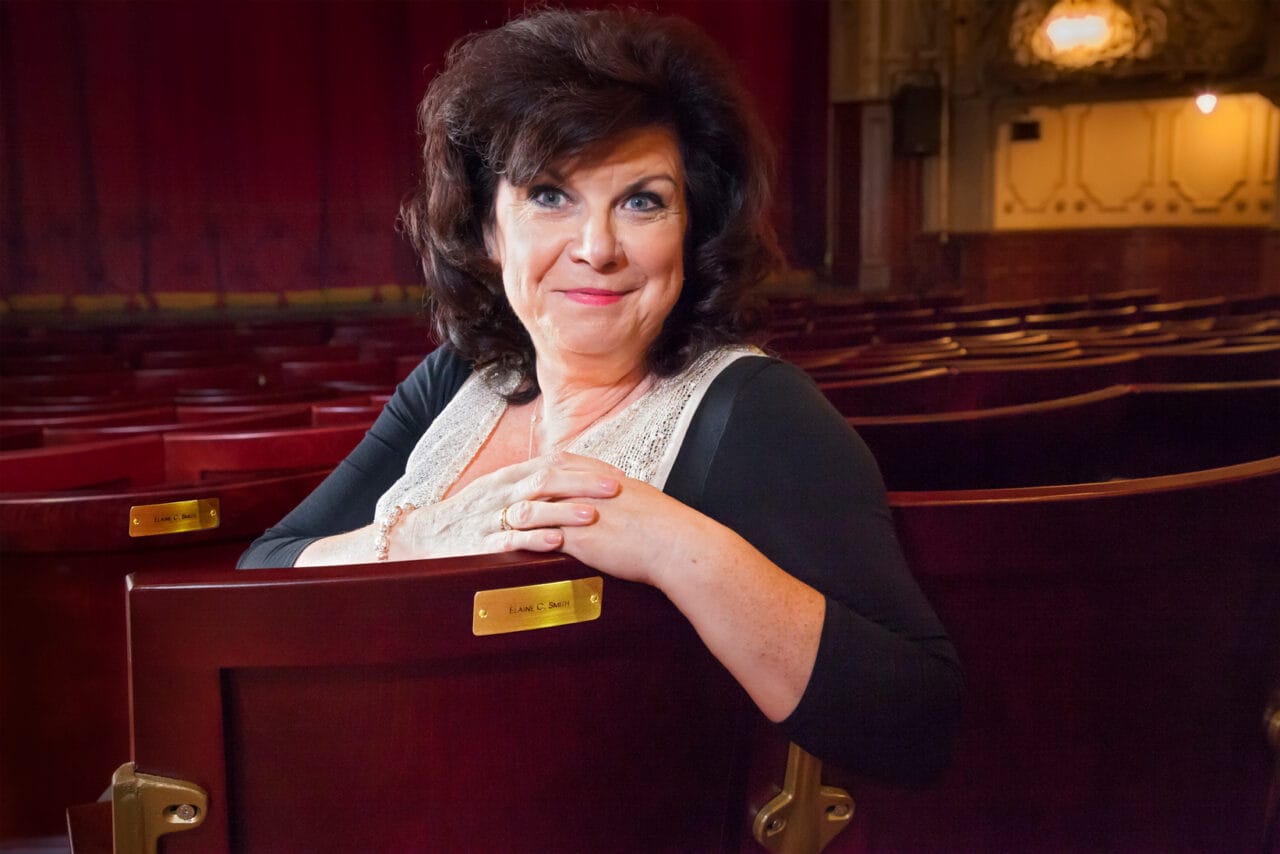 Elaine C Smith, with dark hair and a black top sits and smiles in an empty theatre, resting her arms on the back of a red seat.