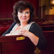 Elaine C Smith, with dark hair and a black top sits and smiles in an empty theatre, resting her arms on the back of a red seat.