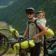 An adult wearing a helmet stands beside a loaded bicycle with a young child in a carrier on their back, surrounded by bikes in a mountainous outdoor setting, showcasing the adventurous spirit of the Red Programme.