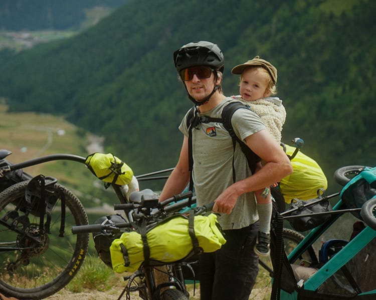 An adult wearing a helmet stands beside a loaded bicycle with a young child in a carrier on their back, surrounded by bikes in a mountainous outdoor setting, showcasing the adventurous spirit of the Red Programme.