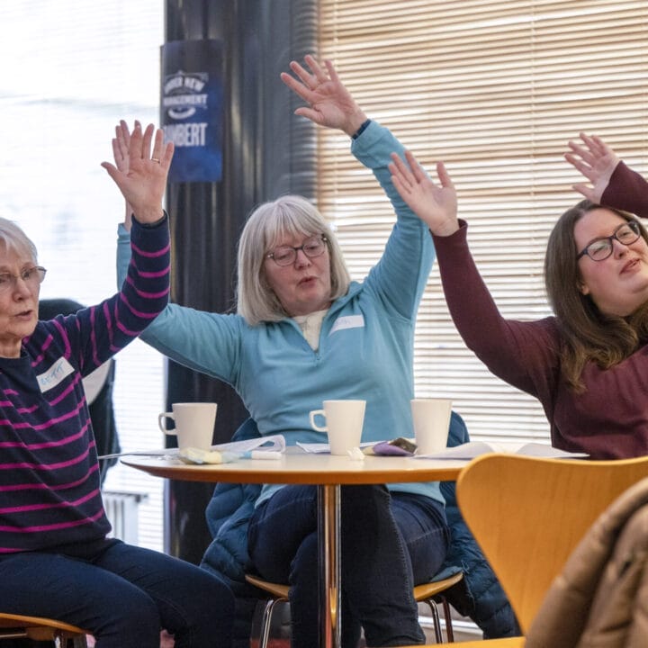 Three women sit at a table with mugs, raising their arms in the air, appearing to participate in a group activity—celebrating being Together in Song in a casual indoor setting.