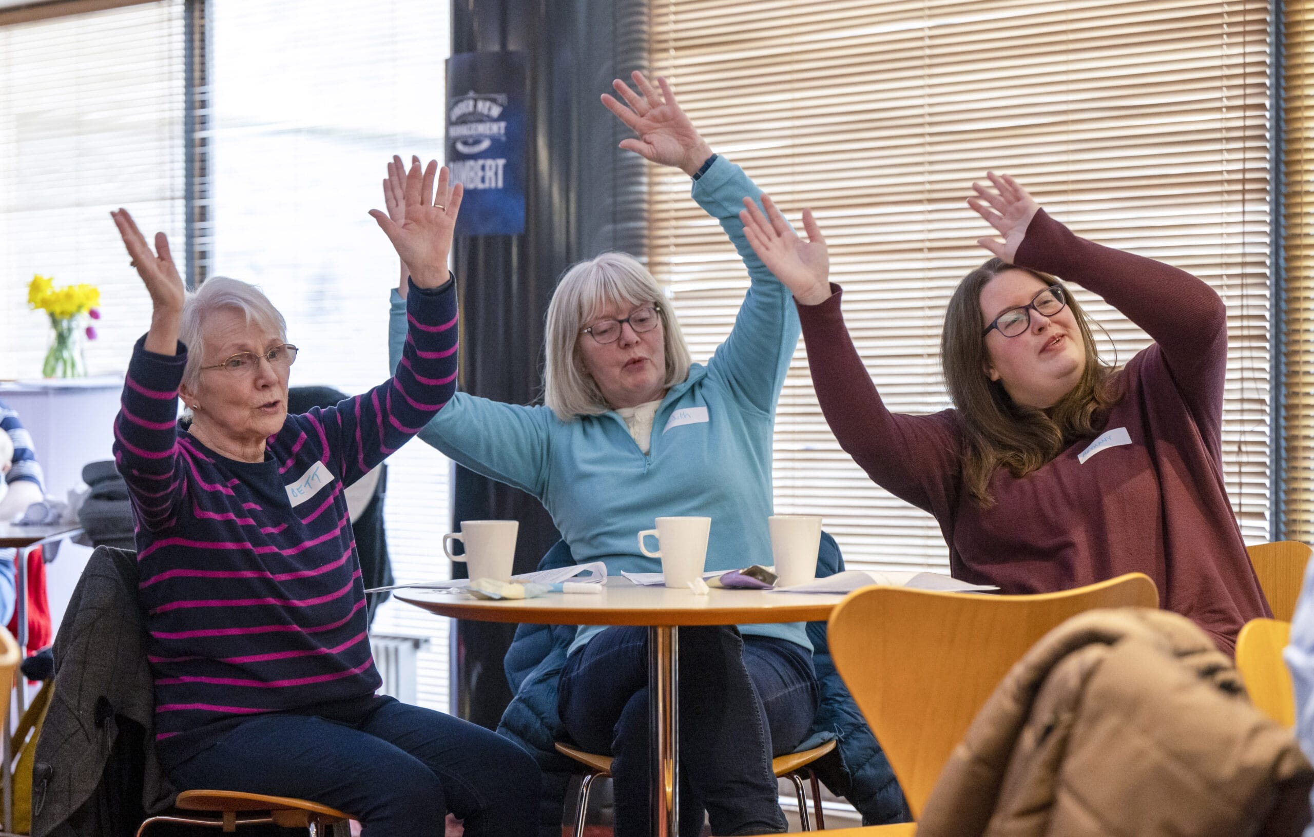 Three women sit at a table with mugs, raising their arms in the air, appearing to participate in a group activity—celebrating being Together in Song in a casual indoor setting.