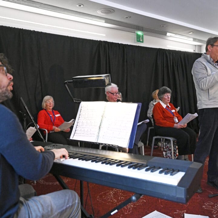 A man sings into a microphone while another plays keyboard; four older adults with sheet music sit Together in Song on a red carpeted stage.