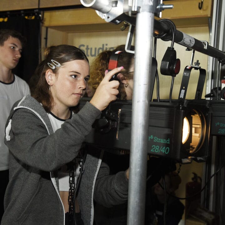 A young woman adjusts stage lighting equipment while a young man and another person observe in the background, gaining valuable Theatre Insights during their time at Holiday Club.