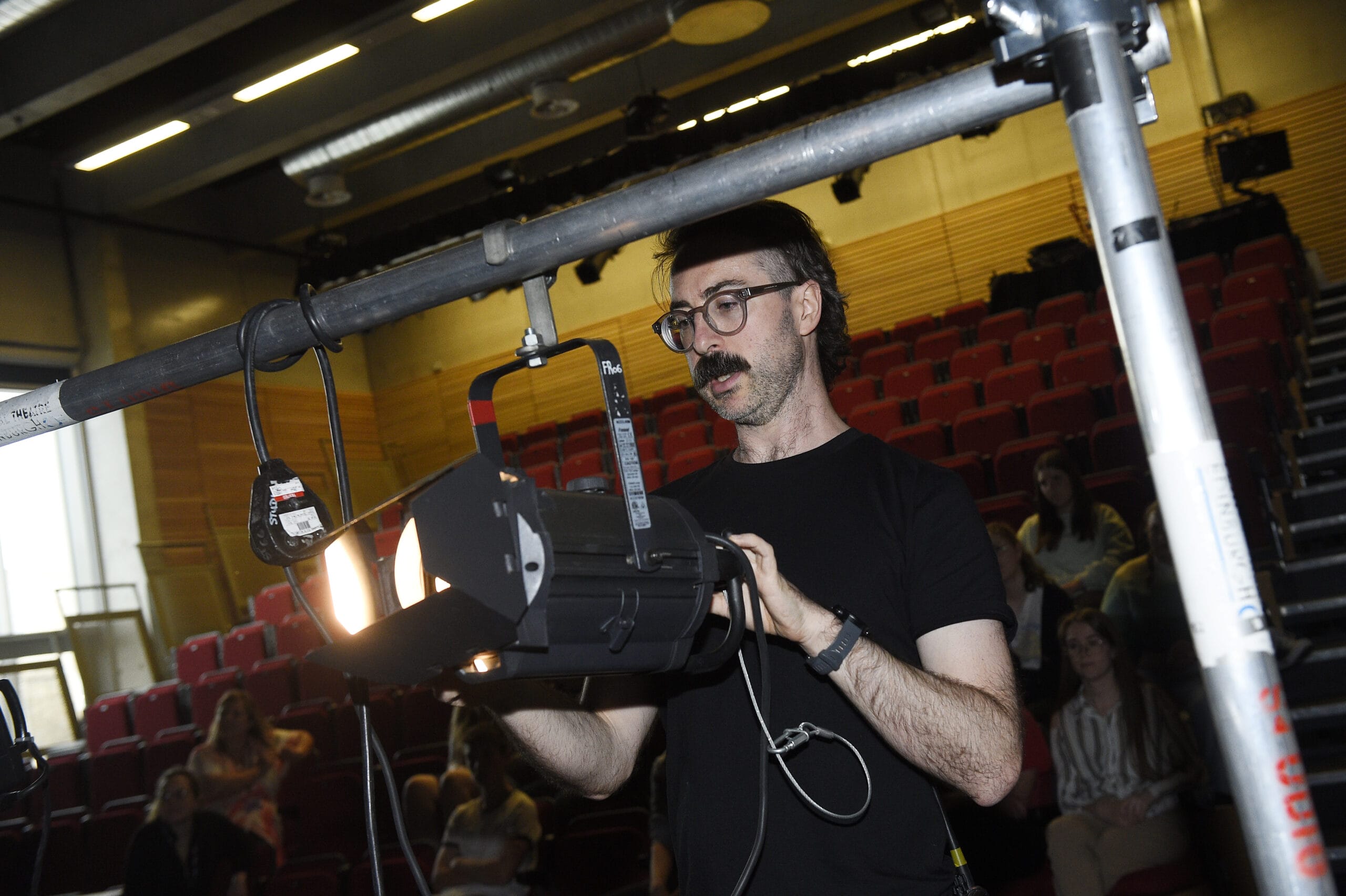 A man adjusts a stage spotlight in a theater with red seats, while a few people observe in the background.