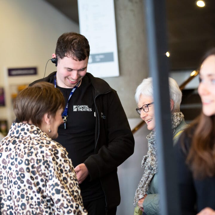 An usher wearing a headset and staff badge smiles while speaking with three women in the theatre foyer.