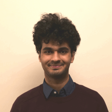A young man with curly dark hair, wearing a dark sweater over a collared shirt, stands against a plain beige background and faces the camera.